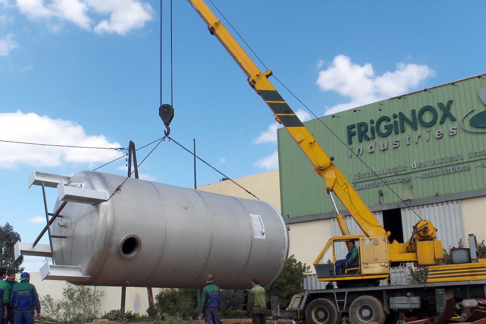 Une capacité en acier inoxydable levée à la grue devant l’atelier Friginox, l’enseigne de l’entreprise en façade