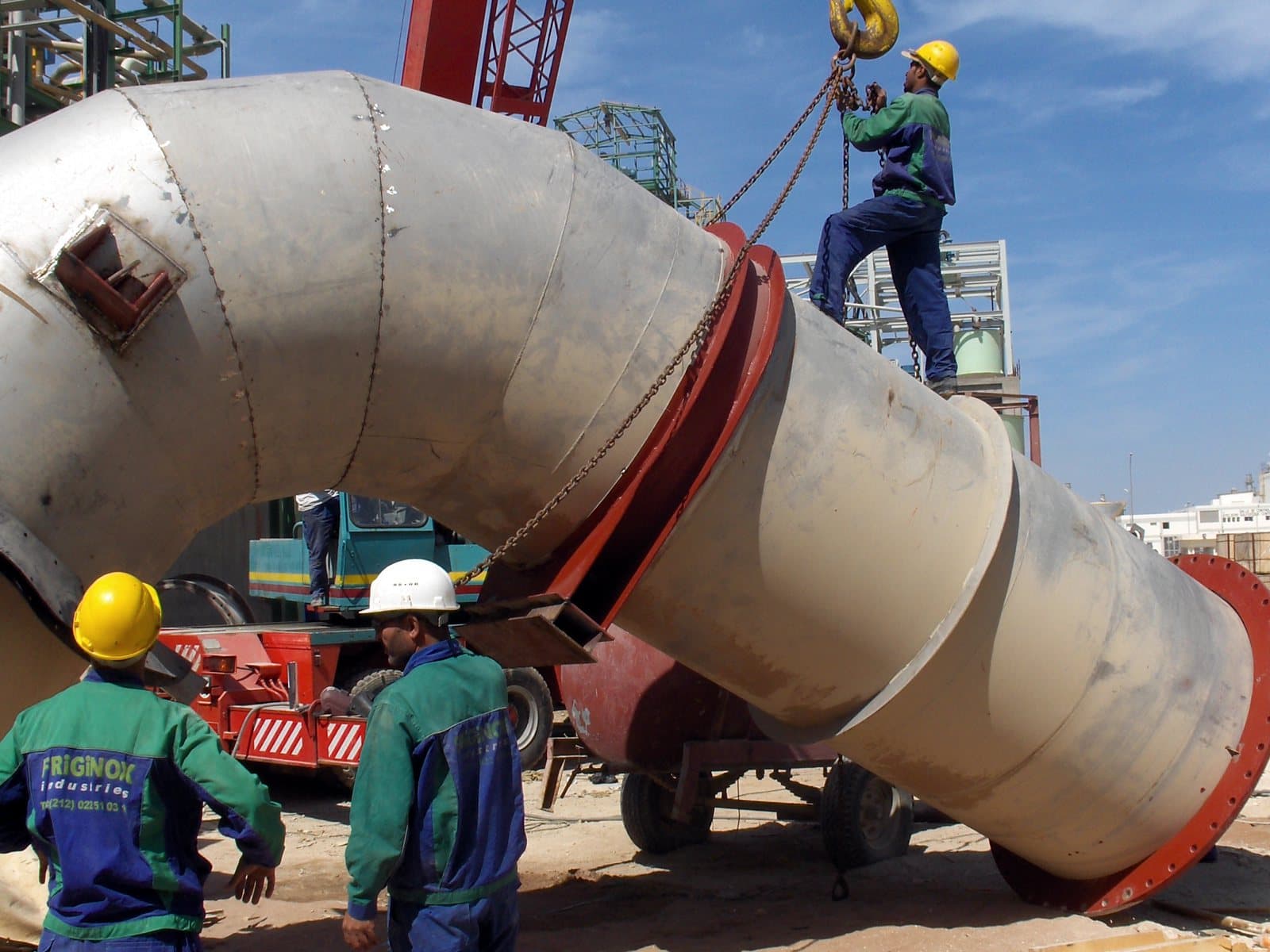 Friginox riggers in company overalls guiding a large steel duct bend slung from a crane on site